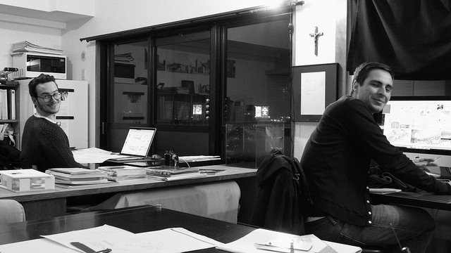 Two people smiling at the camera in the office, with a laptop and papers on the desks (monochrome photo)